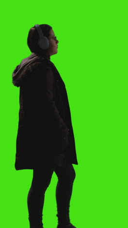 vertical video full length shot of woman wearing wireless headphones streaming music from mobile phone walking across frame against green screen 3