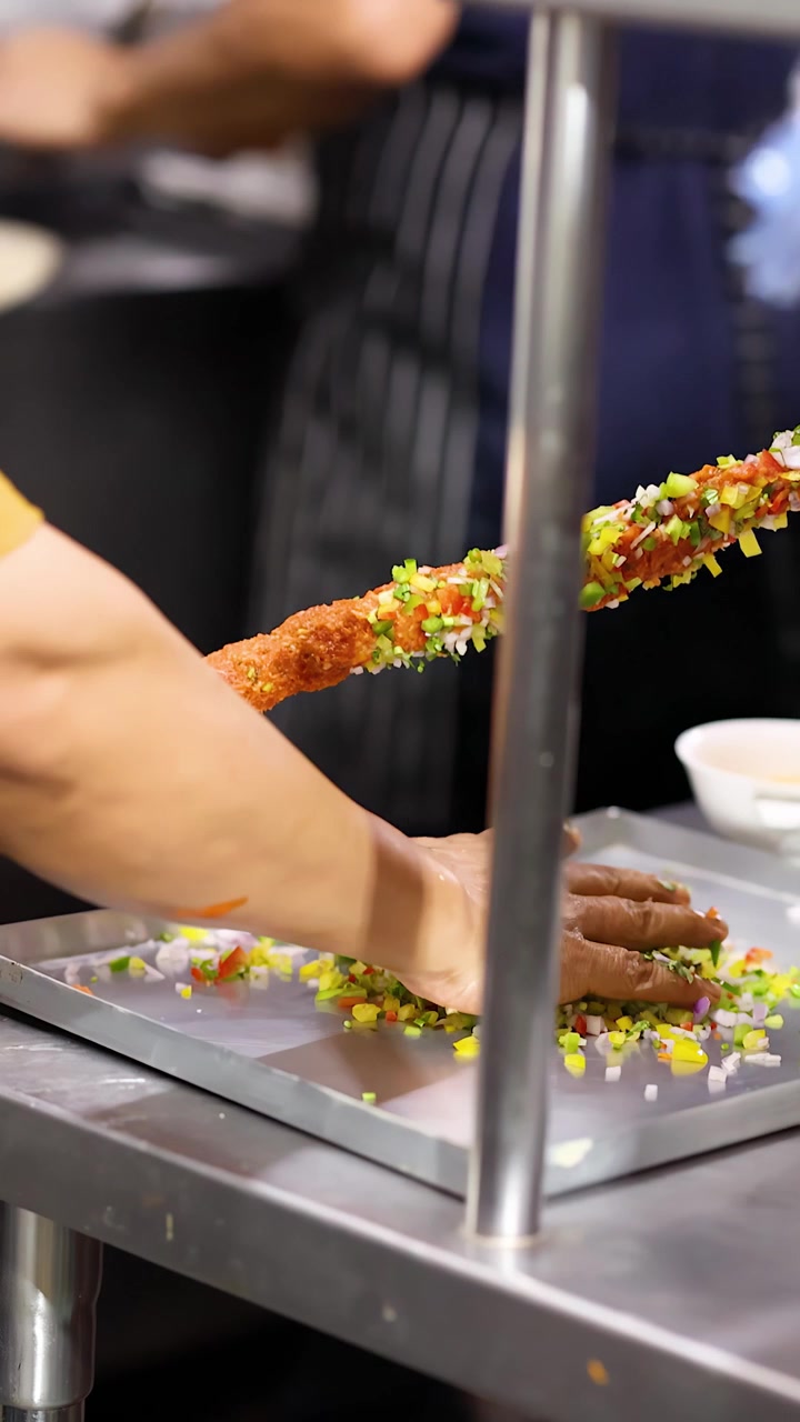 Gros plan d'un chef pressant des brochettes de viande hachée dans des légumes hachés colorés sur un plateau métallique.