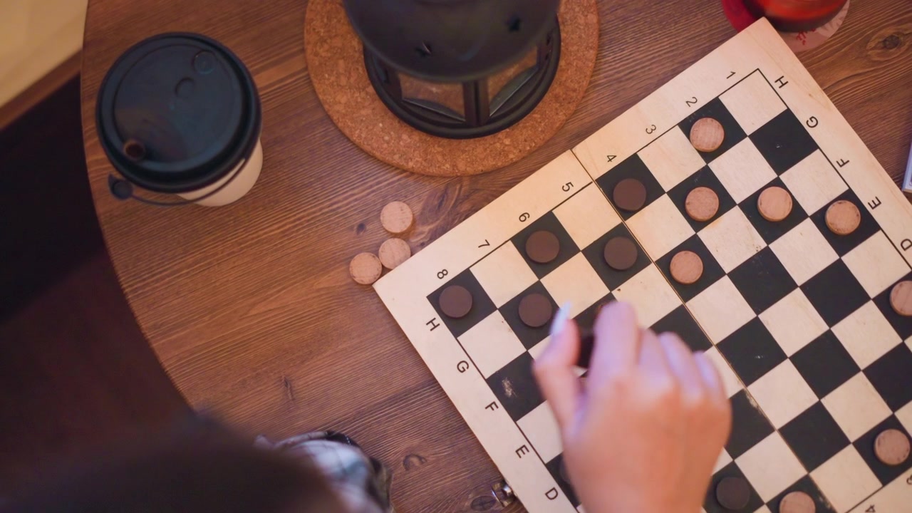 Menina joga damas em mesa de madeira, movendo as peças pelo tabuleiro. Xícara de café e lanterna estão visíveis nas proximidades enquanto a menina se concentra no seu jogo. A cena se passa em ambiente interno com iluminação aconchegante.