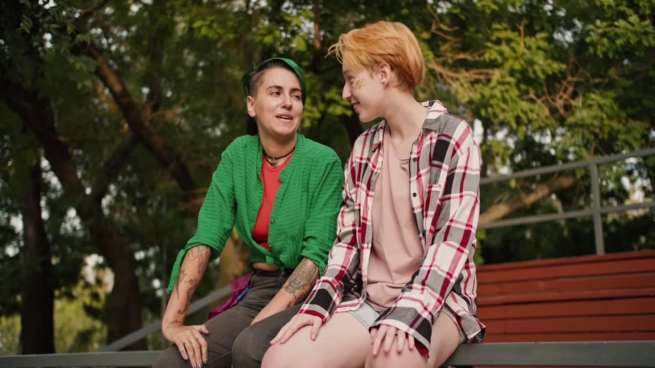 한 lgbt 커플, 크레이드 셔츠를 입은 짧은 머리카락을 가진 도난당한 소녀와 녹색 셔츠을 입는 짧은 녹색 머리카락의 소녀가 공원의 울타리에 앉아 서로 유혹하고 있습니다. 공원에서 데이트를하는 두 레즈비언 소녀.
