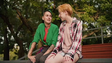 a lgbt couple, a stole girl with short hair in a plaid shirt and a girl with short green hair in a green shirt, are sitting on a fence in the park and flirting with each other. two lesbian girls on a date in the park