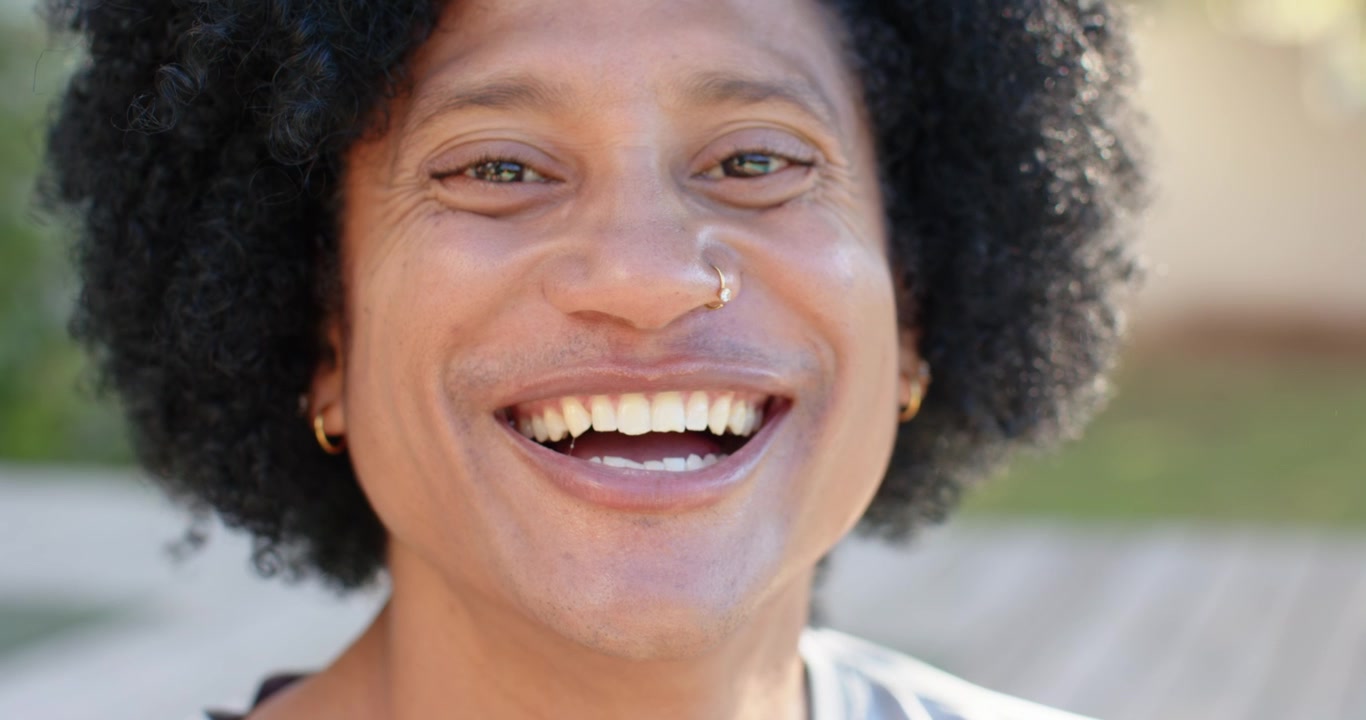 Homem afro-americano reagindo a uma deixa fora da câmera, sorrindo e rindo em um deck ensolarado com uma argola no nariz.