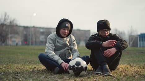 two boys seated on a grassy field, engaged in discussion, with one in a gray top and a soccer ball in front of him, dry grass is scattered around them as they converse