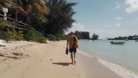 a man walks barefoot along the beach with his shoes in his hands and a satchel over his shoulder