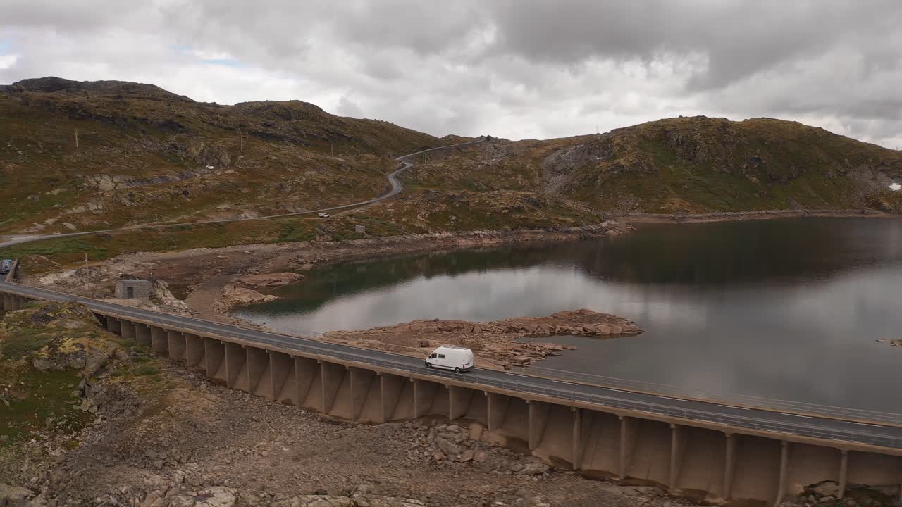 A white van driving across a concrete bridge near a serene lake in Norway's rugged landscape