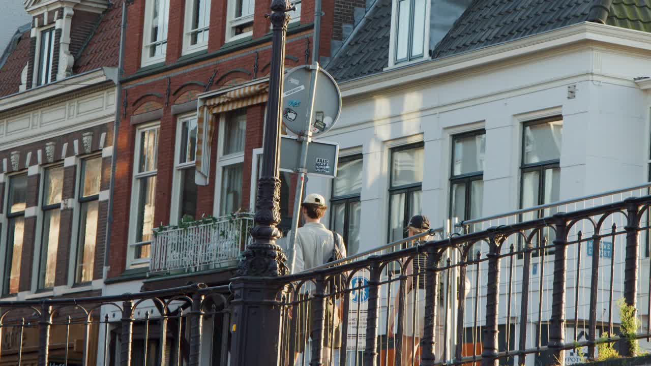 Two individuals cross a classic bascule bridge in Haarlem, Netherlands, with late afternoon sunlight illuminating Dutch canal-side architecture. Static camera, urban atmosphere