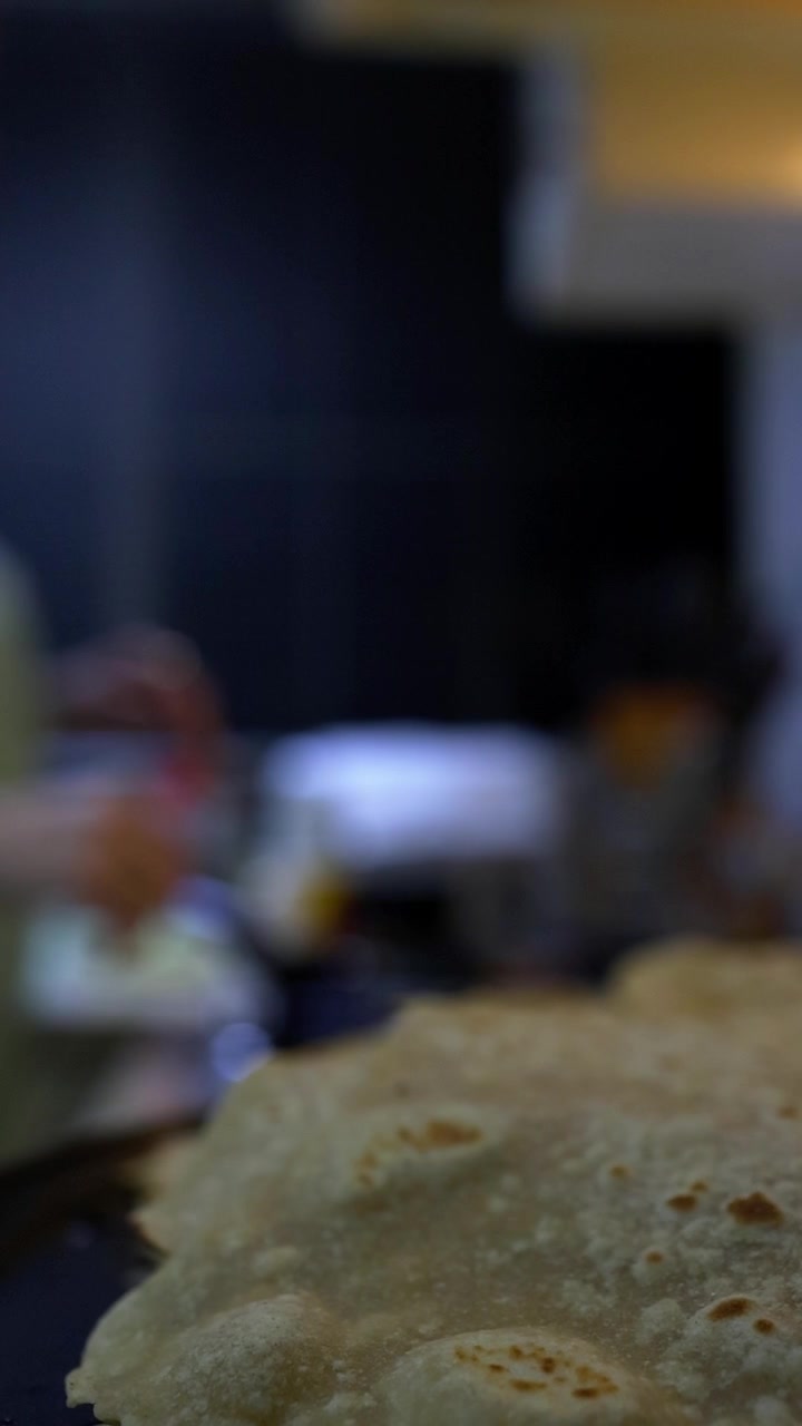 Close-up of freshly cooked chapati with golden-brown spots on a dark plate. A young girl works in a warm, softly lit kitchen preparing traditional Indian flatbread, with blurred background and cooking