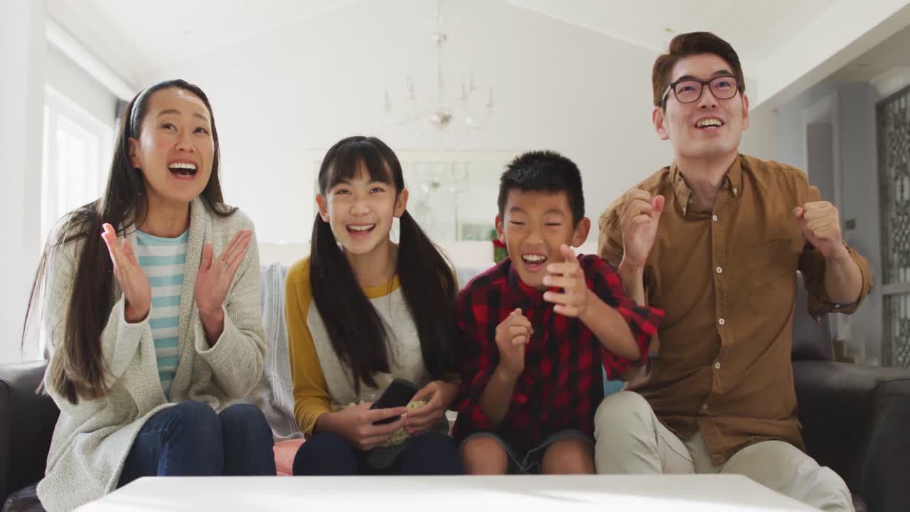 흥분 된 아시아 부모 들 이 tv 에서 스포츠 를 보고 아들 과 딸 과 함께 소파 에 앉아 응원 하고 있다