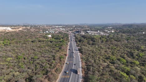 a modern highway cuts through extensive dry woodland, with vehicles heading towards a developing town visible in the distance. the blend of nature and human expansion under a bright, clear sky