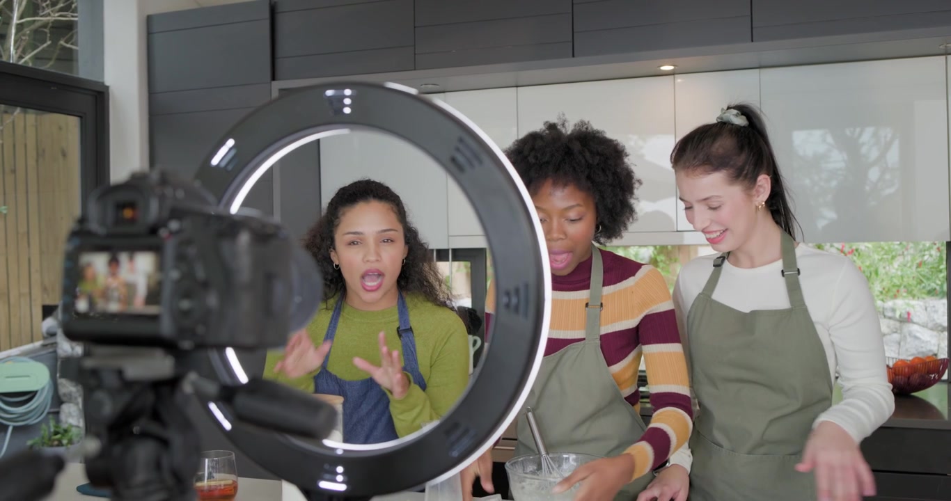 3 women in aprons preparing batter on island with camera filming measuring cracking eggs for demo