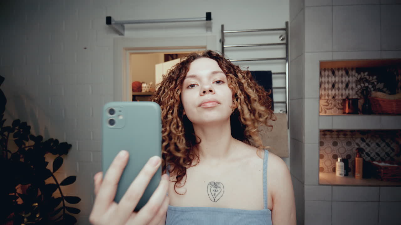 chica de cabello rizado tomando selfies en el espejo con un teléfono inteligente en el baño