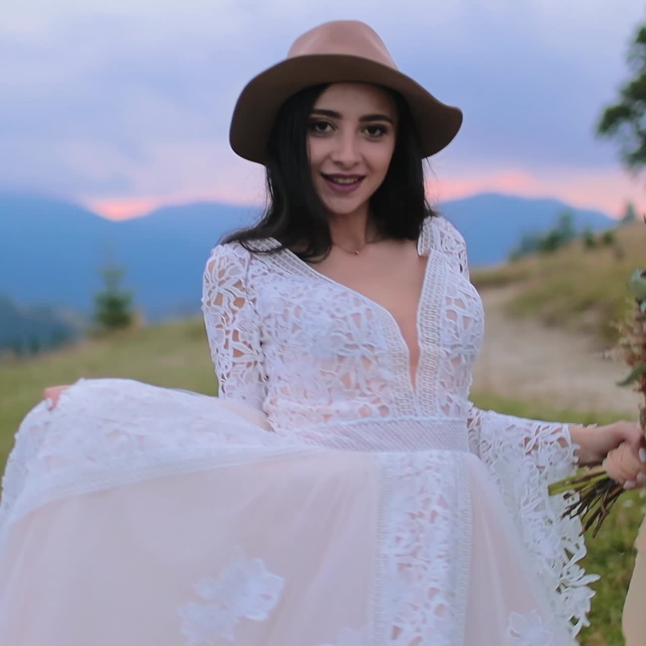 Smiling bride in beautiful white dress. Young woman in hat waving her wedding dress on the nature background in the evening. Girl with bouquet of wild flowers.