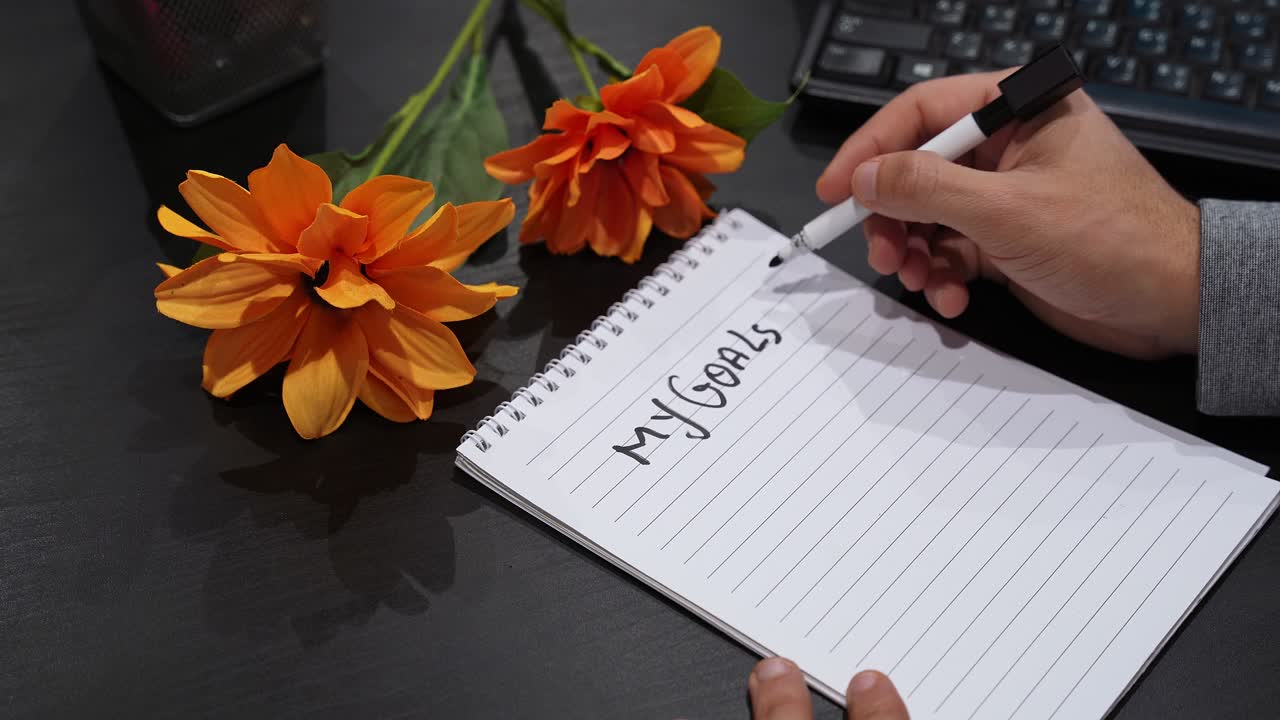 Close-up of hands writing “My Goals 2027” and numbered checklist in spiral notebook, home office desk with laptop keyboard and orange flowers, productivity planning concept with copy space