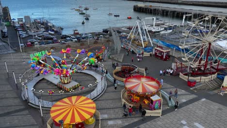 fun fair, dún laoghaire, county dublin, ireland, september 2024. drone orbits clockwise around the fairground rides with the dart station and old ferry terminal in the background.