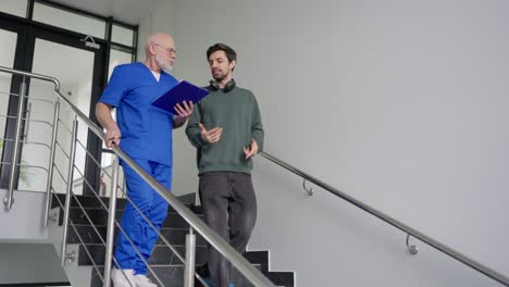 Ein-Junger-Brünetter-Mann-In-Einer-Grünen-Jacke-Und-Kabellosen-Kopfhörern-Kommuniziert-Mit-Einem-Erfahrenen-Arzt-Mit-Grauem-Bart-Und-Blauer-Uniform,-Der-Patientenbeschwerden-In-Sein-Tablet-Schreibt,-Während-Er-In-Einer-Modernen-Klinik-Die-Treppe-Hinuntergeht