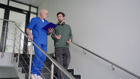 a young brunette guy in a green jacket and wireless headphones communicates with an experienced doctor with a gray beard and a blue uniform who writes patient complaints into his tablet while going down the stairs in a modern clinic