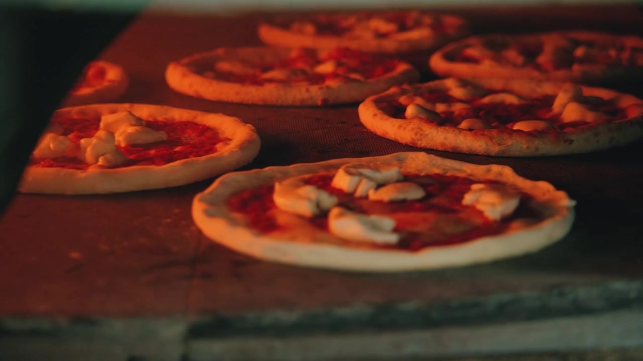 Pizzaer drejer rundt i stenovnen, hvor ilden brænder varmt og kaster et varmt orange lys på pizzaen.