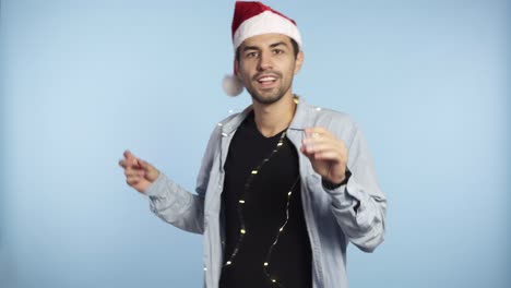 Positive-handsome-man-in-a-Christmas-redhat-and-garland-on-neck-dances-and-smiles-at-the-camera---blue-wall-background-studio