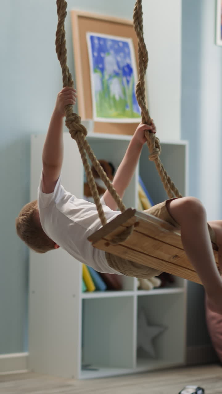 Laughing little boy sits on swing tilting head back. Toddler has fun in light room with bookcase and bean bag chair. Contented kid rests at home