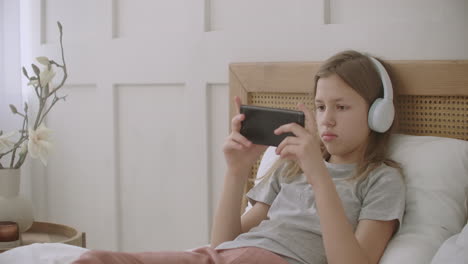 school girl is lying on bed in room and playing game on smartphone by internet listening to music by headphones