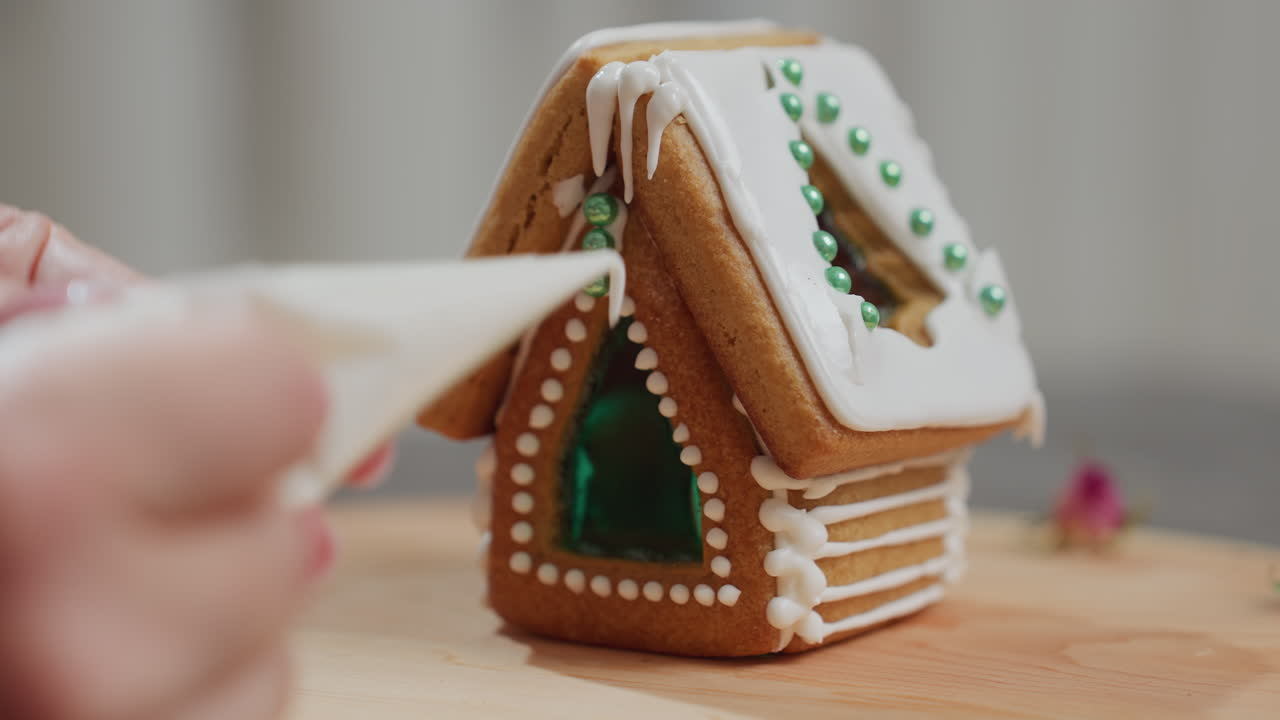 Close-up of hands decorating gingerbread house roof with icing on wooden tray, freshly baked festive cookie structure adorned with green sugar pearls and intricate