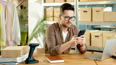 caucasian designer man in eyeglasses sitting at desk texting on mobile phone in clothing shop warehouse