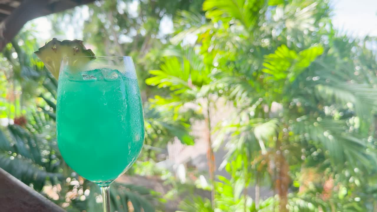 Blue cocktail with pineapple garnish stirred in bright tropical garden, natural daylight, slight camera movement