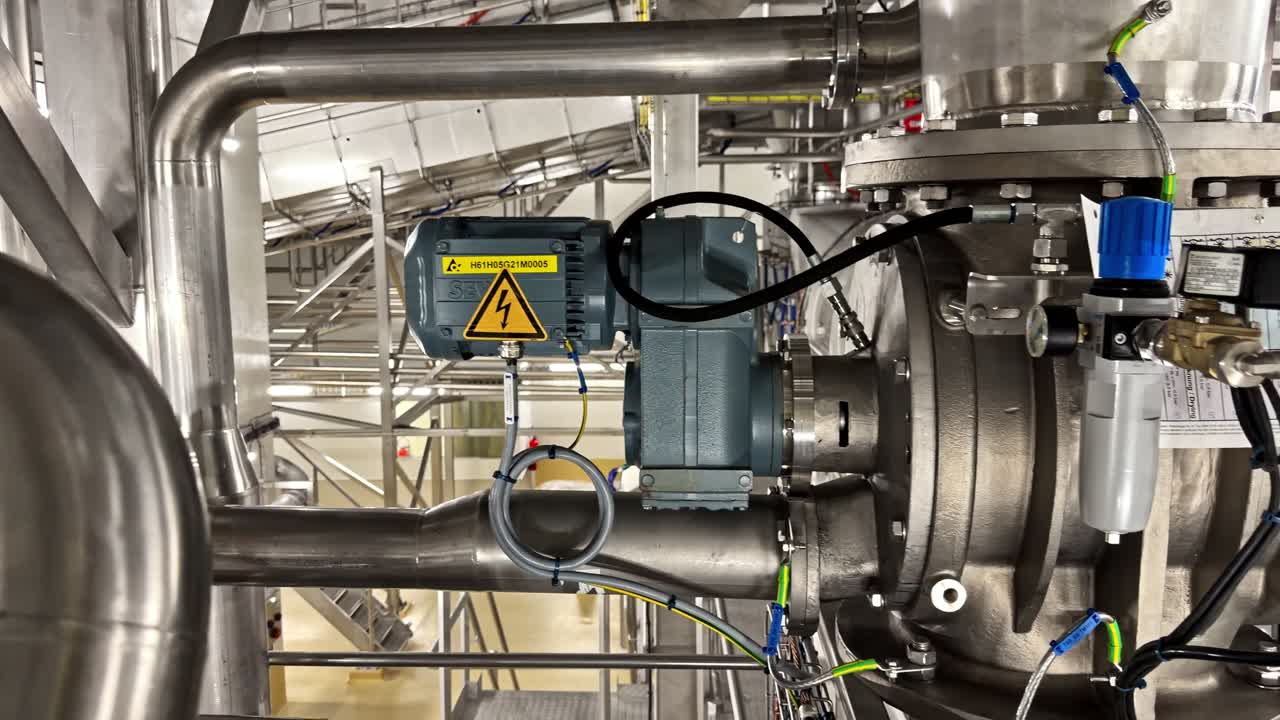 Close-up of stainless steel pipework with motorized valve actuator and high voltage warning in an industrial plant, showing process automation, safety, and modern engineering infrastructure