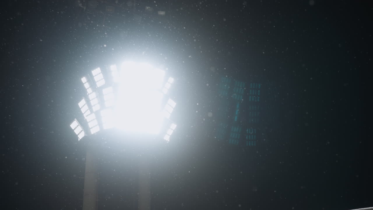 Intense bright stadium light glowing against dark night sky as snowflakes drift in air, creating moody winter atmosphere with light beams scattering through gentle snowfall under cold conditions