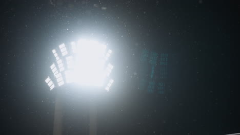 intense bright stadium light glowing against dark night sky as snowflakes drift in air, creating moody winter atmosphere with light beams scattering through gentle snowfall under cold conditions