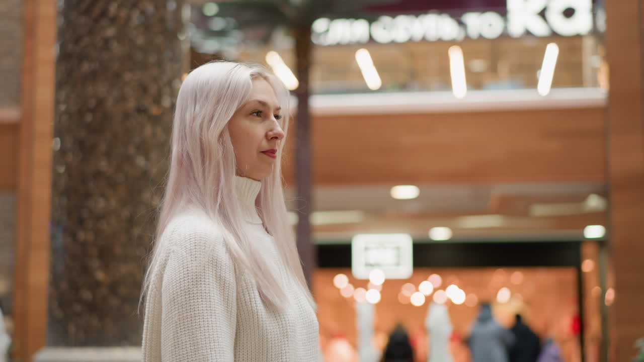 Graceful woman sits beside indoor fountain in shopping mall looks around with smiles ambient shoppers passing under bright ceiling lights over polished tile floor near planters and benches