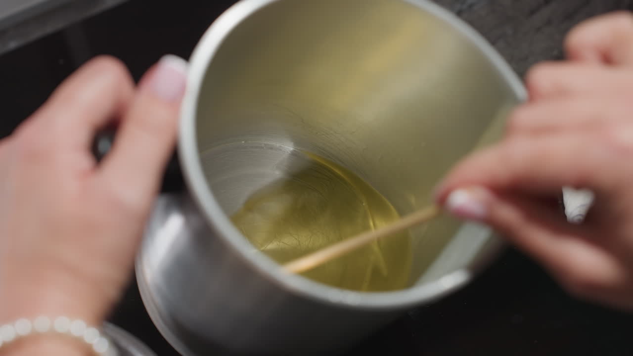 Female baker gently stirs melted butter inside stainless cup using wooden stick in cozy kitchen setup while preparing mixture, showcasing precision and calm focus during baking process
