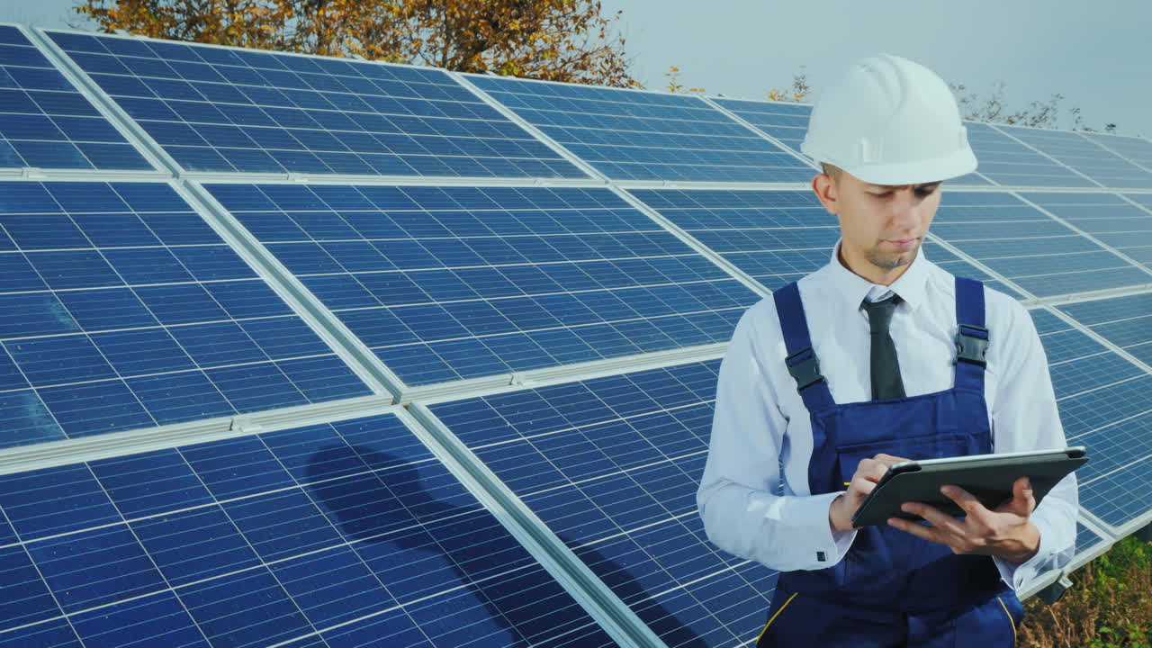 Engineer In Overalls Over A Business Suit Stands Near The Solar Panel Uses A Tablet