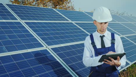 engineer in overalls over a business suit stands near the solar panel uses a tablet