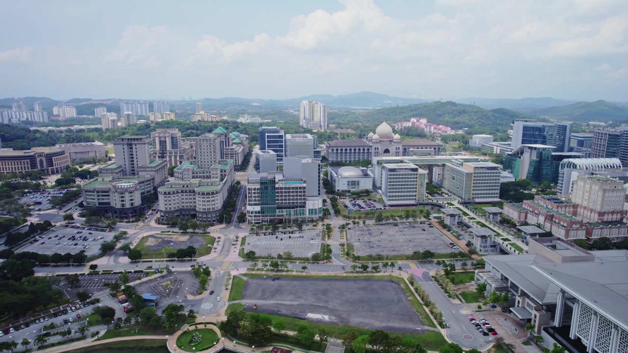 Rekaman udara yang luas menampilkan gedung-gedung pemerintah, kompleks perkantoran, dan perencanaan kota di Putrajaya pada siang hari.