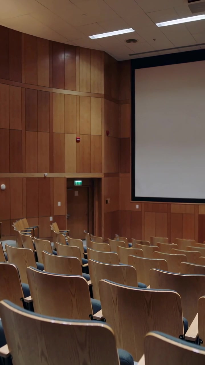Video verticale: Operatore che inizia una panoramica, la telecamera riprende posti a sedere a gradoni e uno schermo in una sala.