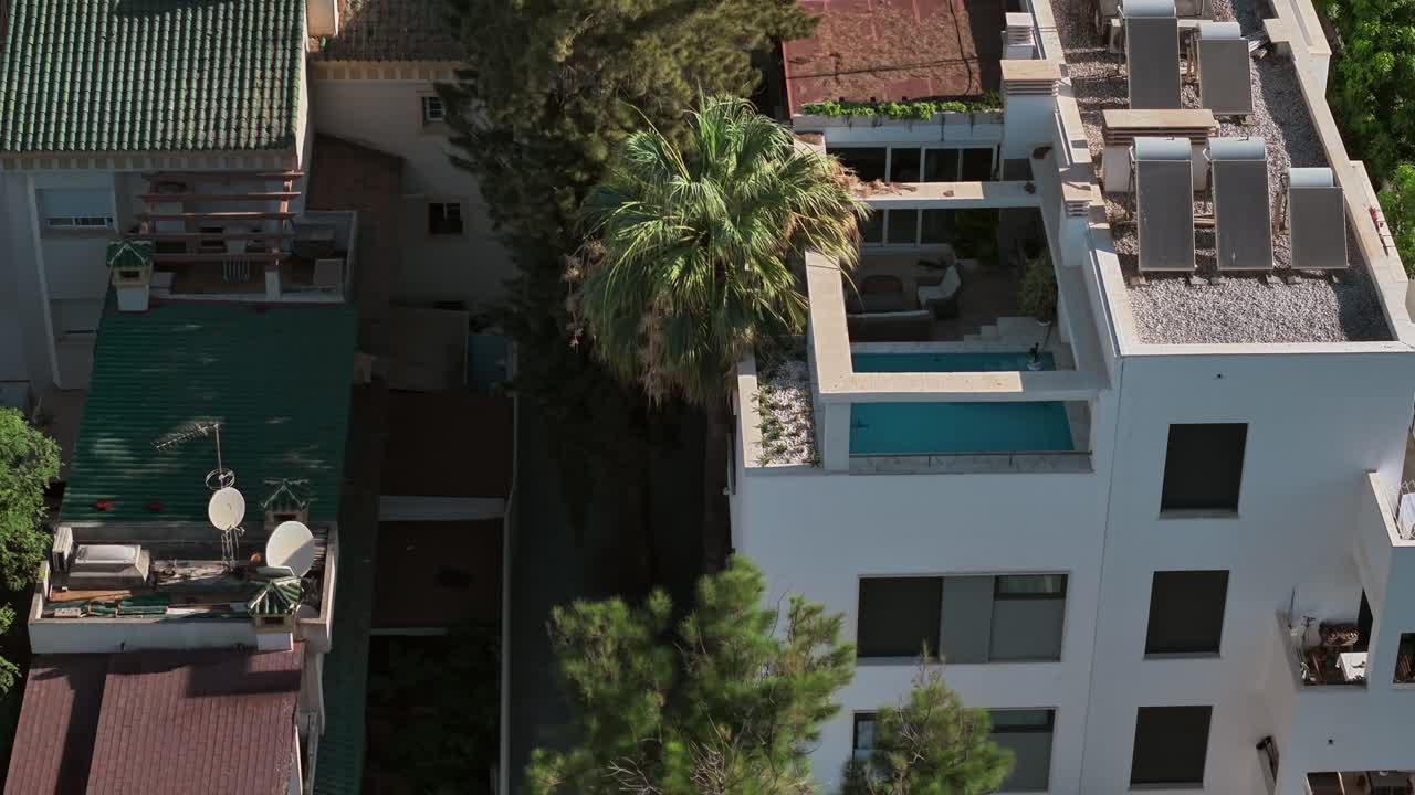 Drone shot captures a modern white residential building in Spain with a rooftop swimming pool. The property also features solar panels, highlighting sustainable and urban lifestyle concepts