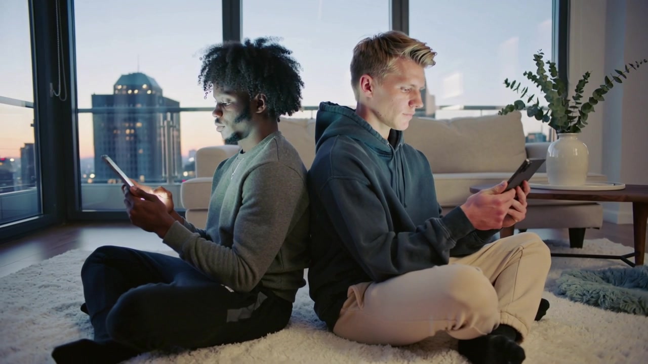 Wearing sweater and hoodie two men sitting back-to-back tapping phones on shag rug at dusk, earbuds