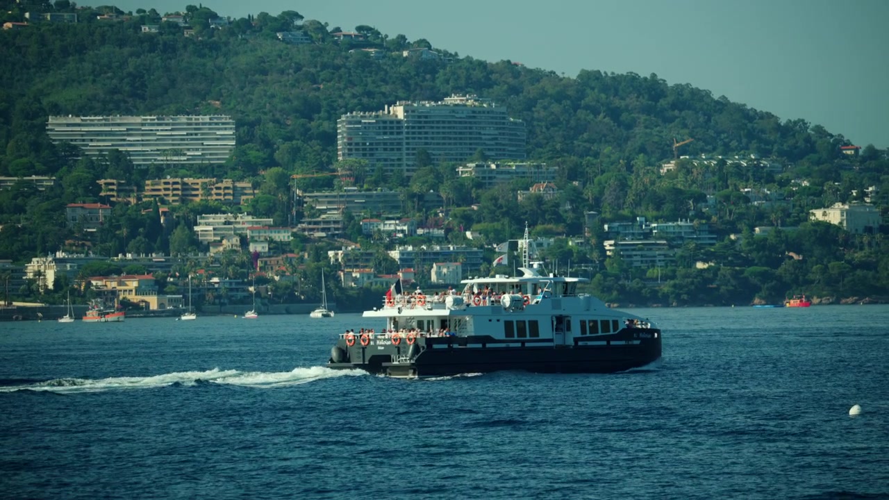 Passenger ferry carrying tourists across the mediterranean sea with the french riviera coastline and lush hills in the background, Cannes, France