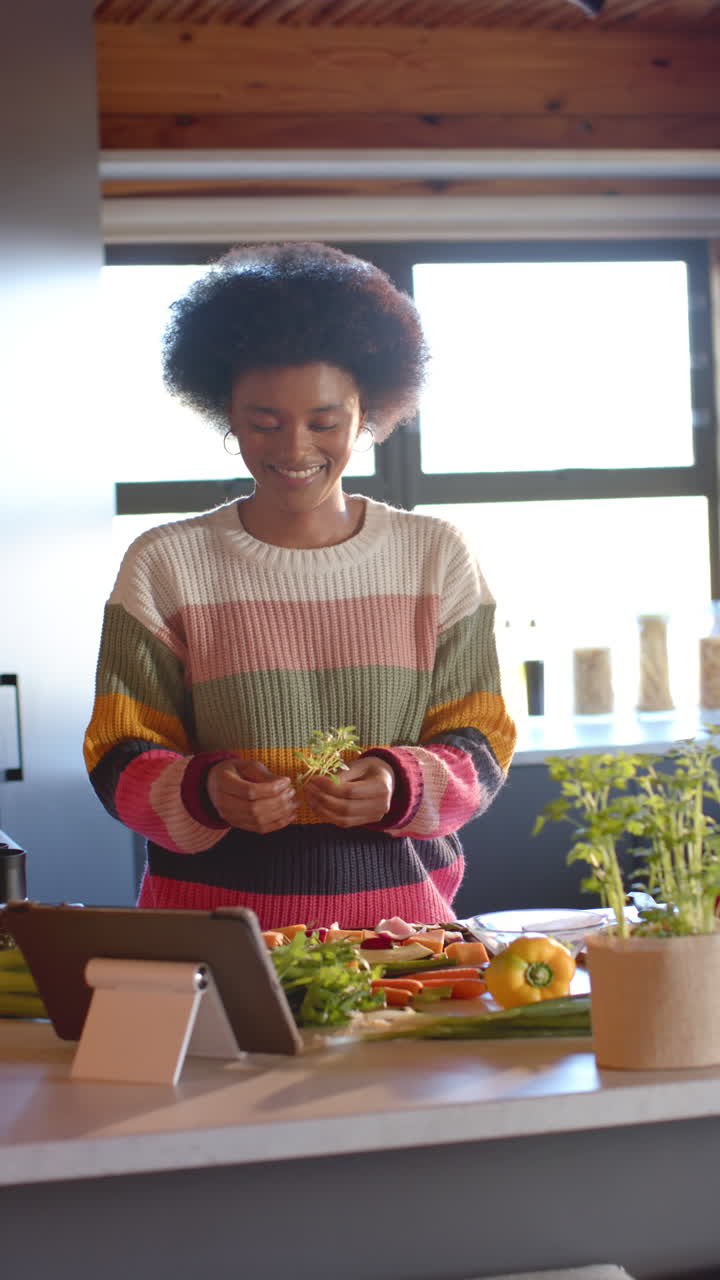 Vertical video of happy african american woman preparing meal using tablet in kitchen, slow motion