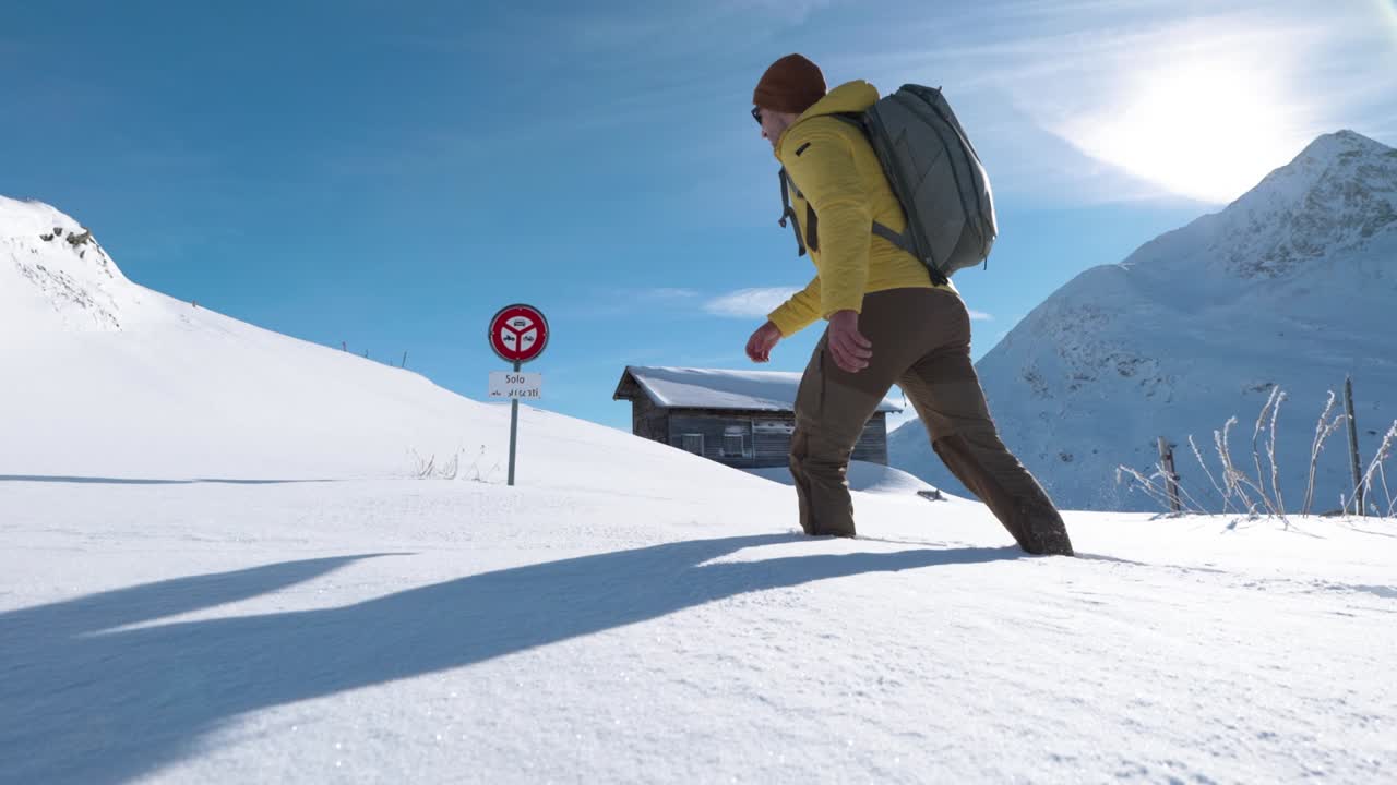 배낭을 메고 노란색 재킷을 입은 백인 청년이 나무 오두막을 배경으로 산에서 맑은 날 깊은 눈 속을 걷는다