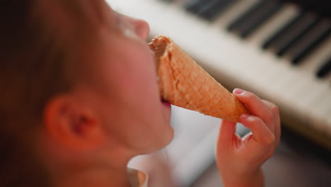 close up overhead view of young girl holding and eating ice cream cone near piano indoors, showcasing sweet and candid moment, soft focus on ice cream and piano keys