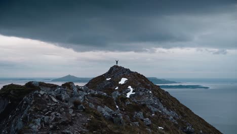 person standing on top of a large mountain with their arms up in the air, surrounded by seascape in norway