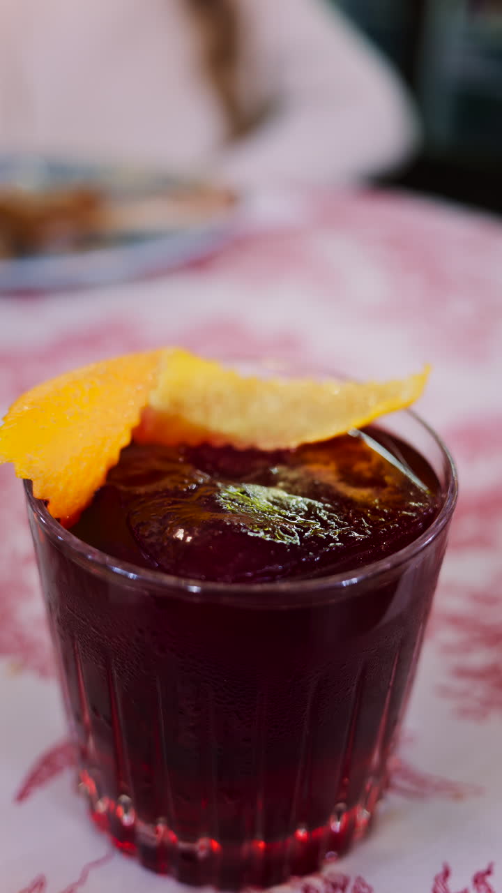 Primo piano di un cocktail Negroni su una tovaglia rossa e bianca, con una donna che beve in un ristorante sullo sfondo. Verticale