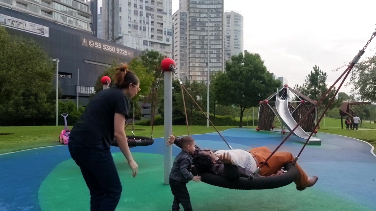 Bayi Dua Tahun Ceria Bersenang-senang di Ayunan Bersama Ibu Muda Latin dan Ayah Berambut Gelap di Taman Bermain Modern. Mereka Berbagi Tawa dan Pelukan, Menikmati Waktu Keluarga Berkualitas