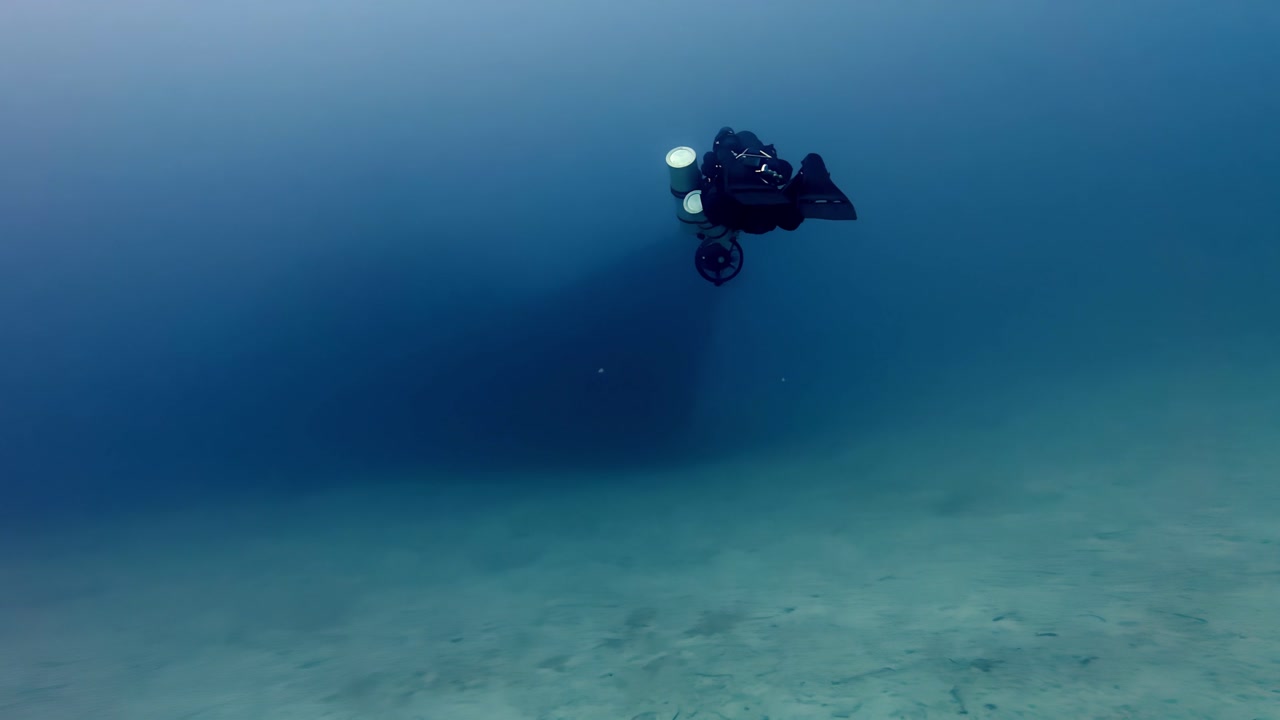 Solo Technical Scuba Diver Exploring Deep Blue Mediterranean Waters Above Sandy Seabed Near Comino Malta During Summer Weather Featuring Dark Equipment Tones and Ethereal Light from a Wide Side Angle