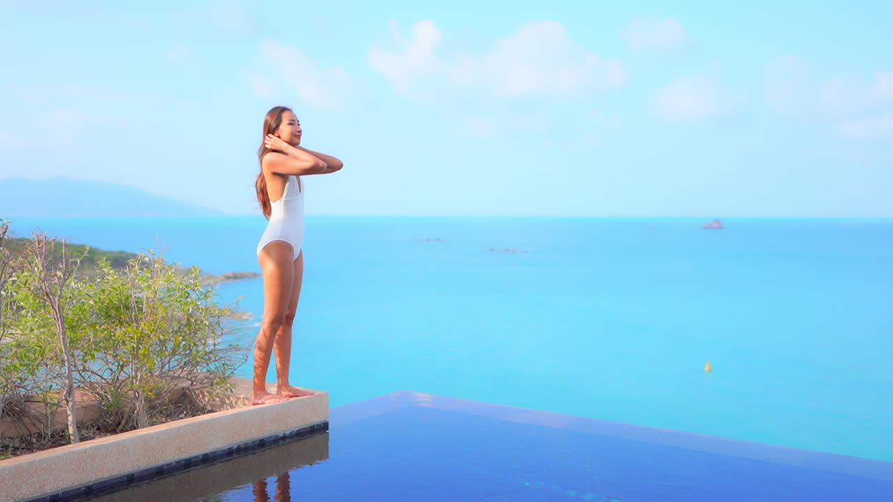 아름다운 섹시한 여성이 바다 지평선을 바라보며 리조트 인피니티 풀의 화분 가장자리에 서 있습니다.