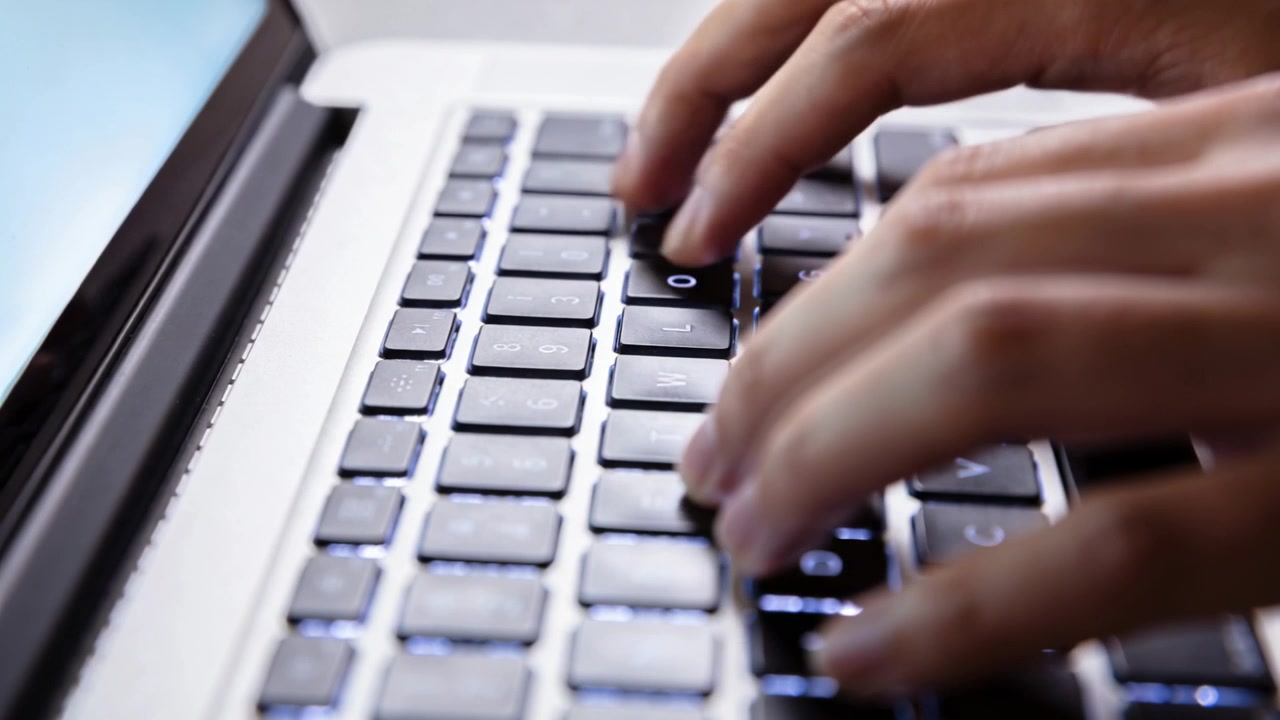 Typing person's hands pressing illuminated keycaps on backlit laptop keyboard at desk, palmrest