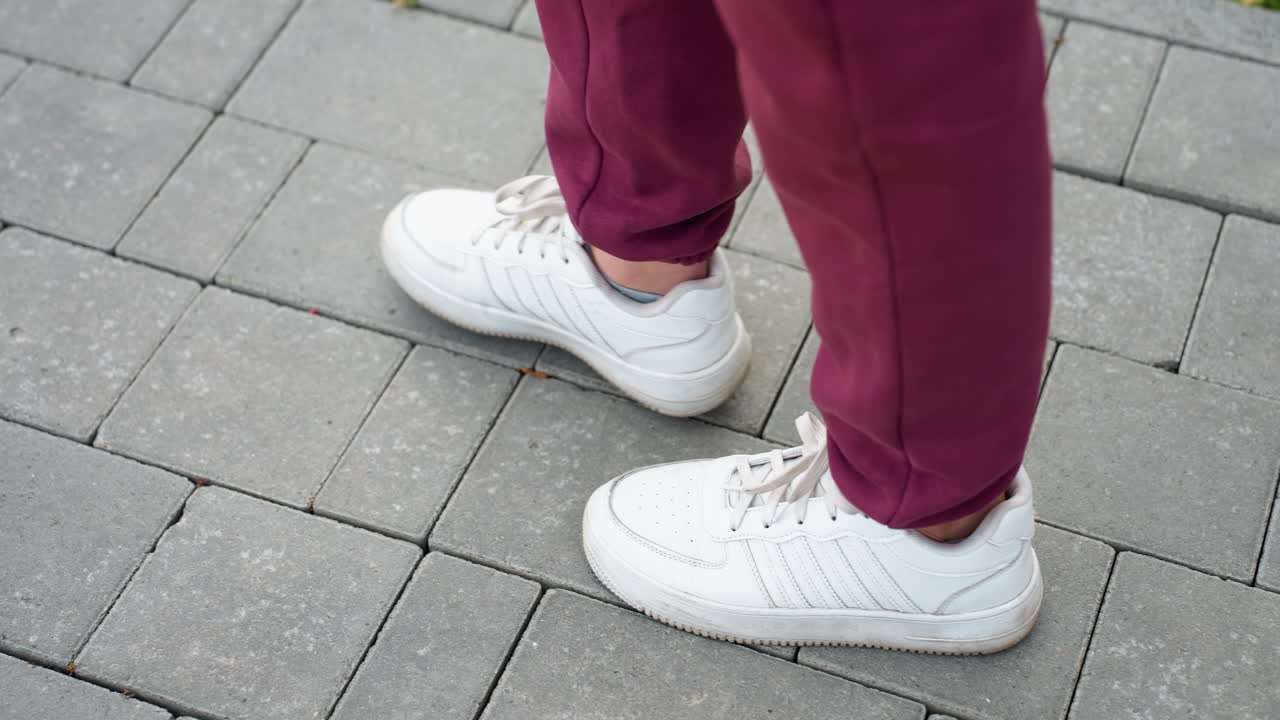 High angle view gym enthusiast in white sneakers and burgundy sports suit walking on gray interlocking tile pavement during outdoor workout session capturing stride movement and urban fitness routine