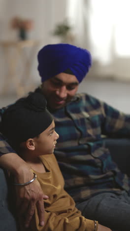 video vertical de un amoroso padre sikh y un hijo joven con turbantes sentados en el sofá en casa hablando juntos 1